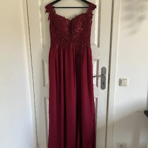 Vinröd långklänning med spetsdetaljer - Elegant vinröd långklänning med spetsdetaljer och hjärtformad urringning. Perfekt för festliga tillfällen med sin glittriga överdel och flytande kjol. Klänningen har breda axelband och en vacker passform.