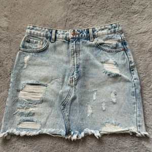 Sliten jeanskjol - Trendig ljusblå jeanskjol med slitna detaljer och fransig nederkant. Kjolen har en klassisk femficksdesign och knäpps med knapp och dragkedja framtill. Perfekt för en avslappnad stil.