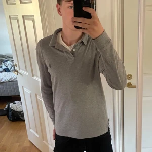 Grå pikétröja från Ralph Lauren - Snygg grå pikétröja från Ralph Lauren med klassisk krage och knappar. Tröjan har långa ärmar och en liten broderad logotyp på bröstet. Perfekt för en stilren look.