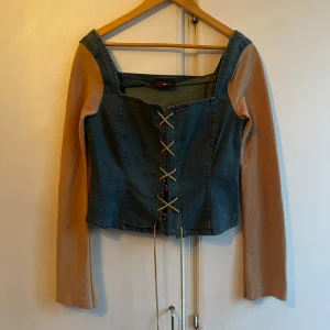 Jeanskorsett med beige ärmar  - Snygg jeanskorsett från Crazy World med långa beige ärmar, snörning framtill och dragkedja på sidan. Perfekt för en unik och trendig look. Korsetten har en fyrkantig urringning och är tillverkad i jeansmaterial.