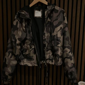 Camouflage vindjacka från Bershka - Snygg camouflage vindjacka från Bershka med dragkedja och justerbar midja. Perfekt för en cool och avslappnad stil. Jackan har långa ärmar och en praktisk huva.