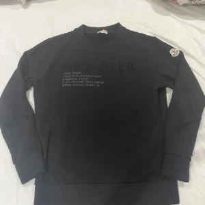 Svart långärmad tröja från Moncler - Snygg svart långärmad tröja från Moncler med tryck på framsidan och ett broderat märke på ärmen. Perfekt för en stilren och bekväm look.