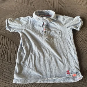 Ljusblå randig pikétröja med ankardetalj - Säljer en ljusblå pikétröja med smala ränder och en liten röd ankardetalj vid nederkanten. Tröjan har korta ärmar, en bröstficka och knappar framtill. Perfekt för en avslappnad stil.