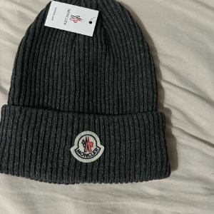 Grå ribbad mössa från Moncler - Snygg grå ribbad mössa från Moncler med broderad logotyp framtill. Perfekt för kyliga dagar och ger en stilren look. Passar både till vardags och mer uppklädda tillfällen.