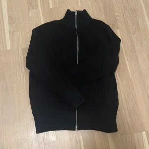 Zip up Maison margiela  - Snygg svart ribbad tröja från Maison Margiela med dragkedja framtill. Perfekt för en stilren look. Tröjan har långa ärmar och en hög krage. Säljer den billigt för har ingen kvitto. 