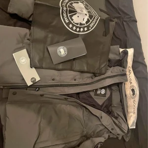 Svart dunjacka från Canada Goose - Säljer en stilren svart dunjacka från Canada Goose med avtagbar pälskrage. Jackan har en praktisk dragkedja och knappar framtill samt en ikonisk logotyp på ärmen. Perfekt för kalla vinterdagar.