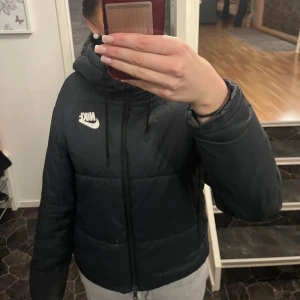 Svart dunjacka från Nike - Säljer en svart dunjacka från Nike med dragkedja och huva. Nike märket har tyvärr börjat lossna lite, då jackan används flitigt men inget man tänker på så mycket.