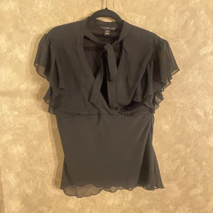 Svart blus från H&M - Elegant svart blus från H&M med volangdetaljer och knytning vid halsen. Blusen har korta ärmar och en lätt transparent design som ger en stilren look. Perfekt för både vardag och speciella tillfällen. Det är storlek 44 men passar mig som oftast har 36/38😚