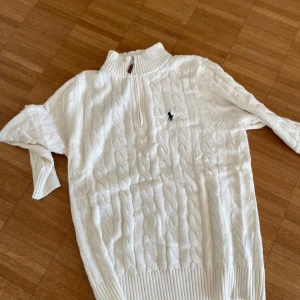 Vit stickad tröja från Ralph Lauren - Säljer en stilren vit stickad tröja från Ralph Lauren med kabelstickat mönster och dragkedja vid halsen. Tröjan har en broderad logotyp på bröstet och ribbade muddar. Perfekt för kyliga dagar! Storlekar: S, M, L, XL och XXL