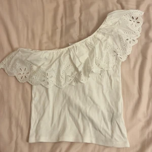 Vit oneshoulder topp från Zara - Säljer en söt vit oneshoulder topp från Zara med vacker spetsdetalj och volang. Aldrig använt! 