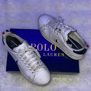 Vita sneakers från Polo Ralph Lauren - Snygga vita sneakers från Polo Ralph Lauren med klassisk logga på sidan. Skorna har snörning och detaljer i rött och blått på hälen. Perfekta för en stilren look. Box medföljer‼️