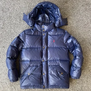 Blå dunjacka från Ralph Lauren - Säljer en snygg blå dunjacka från Ralph Lauren med huva och knappar. Jackan har en quiltad design och är perfekt för kyligare väder. Den har långa ärmar och praktiska fickor med knappar.