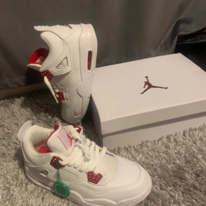 Vita och röda Nike Air Jordans 4 - Snygga vita Nike Air Jordans med röda detaljer. Skorna har snörning och en ikonisk Jumpman-logga på hälen. Perfekta för sneaker-entusiaster som vill ha en stilren look.