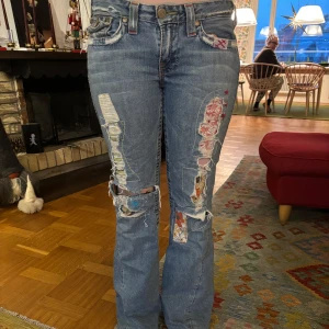 Blå jeans med patchwork och slitningar - Unika blå jeans med coola patchwork-detaljer och slitningar. Byxorna har en bootcut-stil och är lågmidjade. Perfekta för en avslappnad och trendig look.