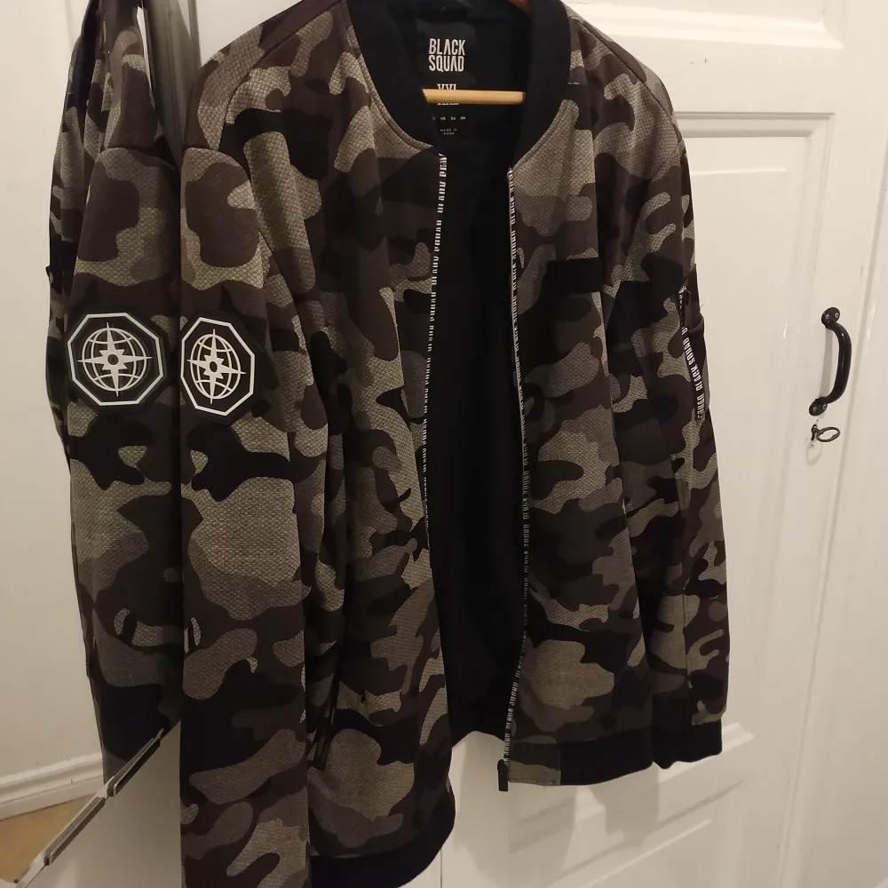 Säljer en snygg camouflage bomberjacka från Black Squad. Jackan har en klassisk bomberstil med dragkedja och ribbade muddar. Den har ett coolt mönster och tryck på ärmarna. Perfekt för en avslappnad stil.. Takit.