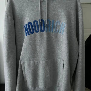 Grå hoodie från Hoodrich - Säljer en stilren grå hoodie från Hoodrich med blå broderad text på framsidan. Den har en klassisk känguruficka och justerbar huva med dragsko. Perfekt för en avslappnad stil.