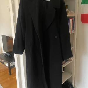 Svart kappa från Weekday - Snygg svart kappa från Weekday med klassisk design. Perfekt för kyligare dagar med sin långa passform och stilrena look. Ett måste i garderoben för den som gillar tidlös stil.