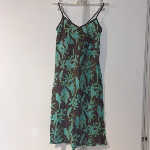 Grönmönstrad klänning - Klänning med turkost bladmönster på en brun/grå bas. Den har smala axelband och en v-ringad design. Lappen är avklippt men ungefär en xs-s i storleken.
