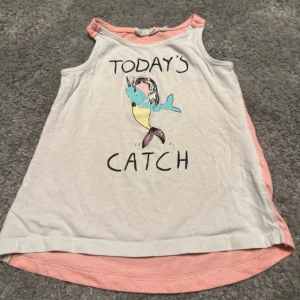 Vit och rosa ärmlös topp med tryck - Söt ärmlös topp i vitt med rosa detaljer och texten 'Today's Catch' på framsidan. Toppen har ett lekfullt tryck av en sjöjungfru och är perfekt för varma sommardagar.