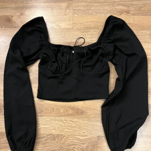 Svart långärmad topp från Nelly - Snygg svart topp från Nelly med långa puffärmar och en smickrande fyrkantig urringning. Toppen har en knytning framtill och är perfekt för en trendig look.