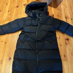 Svart pufferjacka - Säljer en svart pufferjacka med huva och dragkedja. Jackan är tillverkad i 100% polyester och har en långärmad design. Perfekt för kalla dagar.