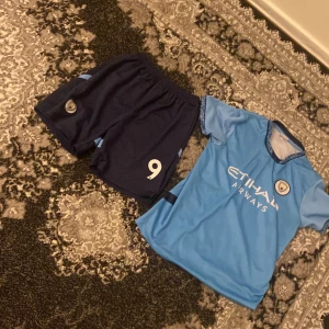 Ljusblå fotbollströja och shorts - Säljer en ljusblå fotbollströja med korta ärmar och matchande mörkblå shorts. Tröjan har texten 'HAALAND' och nummer 9 på ryggen. Perfekt för fotbollsfans!