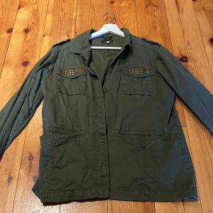 Olivgrön jacka från KappAhl - Snygg olivgrön jacka från KappAhl med coola nitar på bröstfickorna. Jackan har långa ärmar och knäppning fram. Perfekt för en avslappnad stil med en touch av edge.