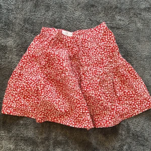 Röd blommig kjol - Charmig röd kjol med ett sött blommönster i vitt. Kjolen har en elastisk midja för bekväm passform och är perfekt för varma dagar.