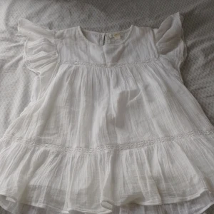 Vit blus med volangdetaljer - Charmig vit blus med volangdetaljer på ärmarna och nederkanten. Blusen har en luftig passform och är perfekt för varma dagar. Den är tillverkad i ett lätt och skönt material.