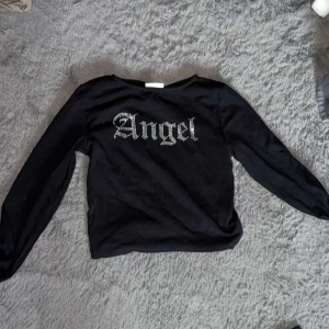 Svart långärmad topp med text - Snygg svart långärmad topp med texten 'Angel' i glittriga bokstäver på framsidan. Perfekt för en cool och avslappnad stil. Passar bra till jeans eller kjol.