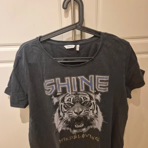 Svart t-shirt med tigertryck från b.young - Cool svart t-shirt från b.young med ett stort tigertryck och texten 'SHINE' i blått och guld. Perfekt för en avslappnad stil med en touch av attityd. XL/L