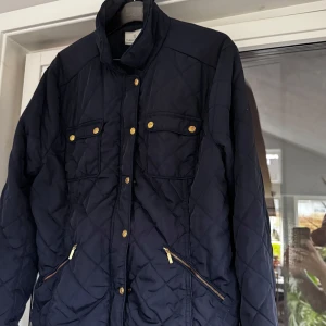 Mörkblå quiltad jacka från Flash Woman - Snygg mörkblå quiltad jacka från Flash Woman med guldknappar och dragkedjor. Jackan har en klassisk krage och två bröstfickor med knappar. Perfekt för kyligare dagar. Kika och se om du ser något mer i min profil som du gillar så skickar jag med det till dig också!😊 Fler kläder kommer! 