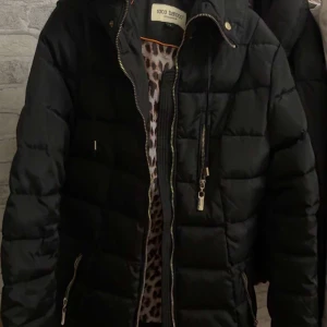 Svart pufferjacka från Roco Baroco - Snygg svart pufferjacka från Roco Baroco med dragkedja och knappar. Jackan har en huva och är perfekt för kyliga dagar. Insidan har ett leopardmönster som ger en cool kontrast. Perfekt för dig som vill hålla stilen i vinter. Den har en svart päls också.