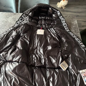 Svart dunväst från Moncler - Snygg svart dunväst från Moncler med dragkedja och huva. Västen har en glansig finish och Moncler-logga på ryggen. Perfekt för kyligare dagar. Köpt för 1350 knappast använd 
