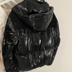 Svart glansig pufferjacka med huva - Snygg svart pufferjacka med glansig yta!! Jackan har huva och stängs med både dragkedja och knappar. Perfekt för kyliga dagar🩷🩷storlek M men sitter perfekt på mig som brukar ha xs/s🩷kan gå ner i pris vid snabb affär!! 