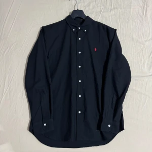 Svart skjorta från Ralph Lauren - Snygg svart skjorta från Ralph Lauren med klassisk krage och knappar framtill. Den har en diskret röd broderad logotyp på bröstet och långa ärmar. Perfekt för en stilren look.