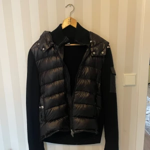 Svart pufferjacka med stickade ärmar från Moncler - Snygg svart pufferjacka från Moncler med stickade ärmar och huva. Jackan har en dragkedja framtill och tryckknappar vid fickorna. Perfekt för kyliga dagar med sin kombination av quiltad kropp och mjuka ärmar. Går att scanna koden ifrån moncler märket vid armen