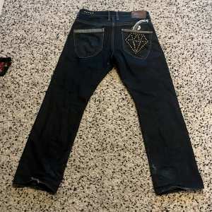 Mörkblå jeans med nitar från Artful Dodger - Snygga mörkblå jeans från Artful Dodger med coola nitdetaljer på bakfickorna och längs sidorna. Jeansen har en straight passform och klassisk femficksdesign. Perfekta för en trendig look!