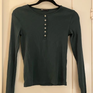 Mörkgrön ribbad tröja från Zara - Säljer en stilren mörkgrön ribbad tröja från Zara med knappar framtill. Tröjan har långa ärmar och en rund halsringning. Perfekt för en avslappnad look.