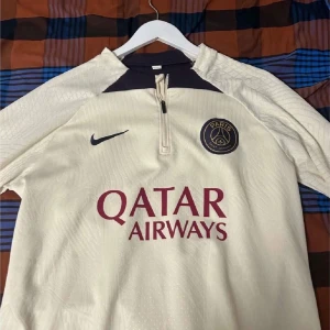 Beige Psg fotbollströja  - Snygg beige fotbollströja från Nike med Paris Saint-Germain-logga och Qatar Airways sponsortryck. Tröjan har en dragkedja vid halsen och korta ärmar. Perfekt för fotbollsfans som vill visa sitt stöd för PSG! Funkar för både storlek S och M. Perfekt inför sommaren.