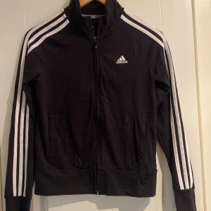 Svart träningsjacka från Adidas - Snygg svart träningsjacka från Adidas med klassiska vita ränder längs ärmarna. Jackan har dragkedja framtill och en hög krage. Perfekt för sport och vardag. Bra skick storlek 36
