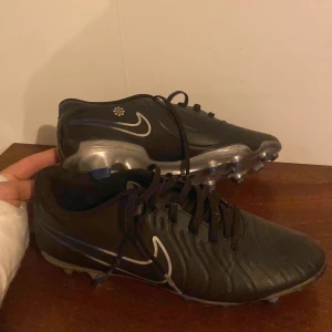 Svarta fotbollsskor från Nike - Säljer ett par svarta fotbollsskor från Nike med vita detaljer och snörning. Skorna har dobbar för bra grepp på planen och en stilren design med silverfärgade inslag. Priset kan diskuteras. Köpta för 750kr