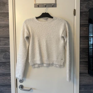 Vit fluffig tröja från Tiffosi - Mysig vit fluffig tröja från Tiffosi i storlek 164 cm. Perfekt för kyliga dagar med sin mjuka och bekväma design. Tröjan har långa ärmar och en rund halsringning.