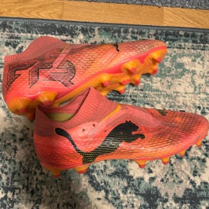  fotbollsskor - Snygga röda fotbollsskor från Puma med svart logotyp och orange detaljer. Skorna har en strumpliknande passform och dobbar för bra grepp på planen. Perfekta för fotbollsspelare som vill ha stil och funktion!  De är den dyraste modellen (Elite)