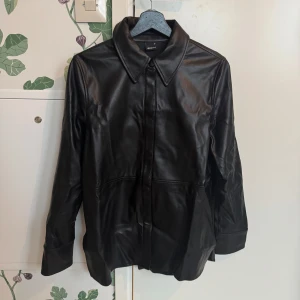 Svart skinnjacka från Gina Tricot - Snygg svart skinnjacka från Gina Tricot med klassisk krage och långa ärmar. Perfekt för en stilren look. Jackan har en slät yta och stängs med knappar framtill.