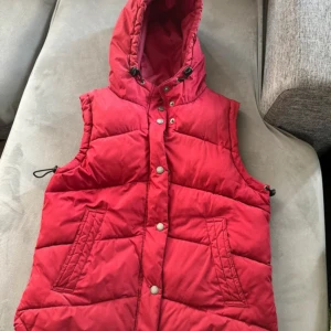 Röd dunväst från North Rock Sports - Säljer en snygg röd dunväst från North Rock Sports. Västen har en praktisk huva och stängs med knappar. Perfekt för kyligare dagar och ger en sportig look.