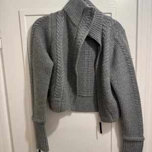 Grå stickad kofta/jacka från Malene Birger - Säljer en stilren grå stickad kofta från Malene Birger. Koftan har en dragkedja framtill och ett snyggt flätmönster. Den är perfekt för kyliga dagar med sin höga krage och långa ärmar. 100% ull . Passar flesta storlekar