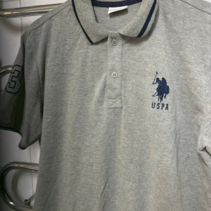 Grå pikétröja från U.S. Polo Assn. - Snygg grå pikétröja från U.S. Polo Assn. med klassisk krage och knappar. Tröjan har en broderad logga på bröstet och kontrastfärgade detaljer på kragen. Tröjan är använd men i bra skick.