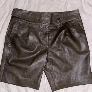 Bruna skinnshorts från Margit Brandt - Snygga bruna skinnshorts från Margit Brandt med hög midja och knappdetalj. Perfekta för en stilren look. De har fickor fram och en bakficka för extra funktionalitet.