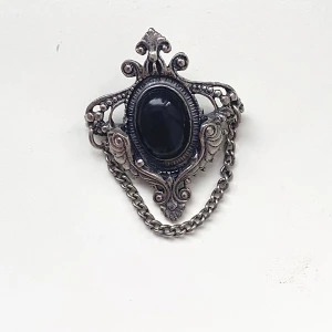 Vintage brosch med svart sten - Elegant gothisk vintage brosch i silverfärgad metall med en stor svart oval sten i mitten. Detaljerad design med snirkliga mönster och en kedja som hänger ned. Perfekt för att ge en klassisk touch till din outfit.